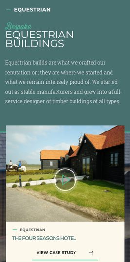 Black wooden buildings with red roofs beside a grassy field and a horse rider, used as an example of timber construction by an equestrian company.