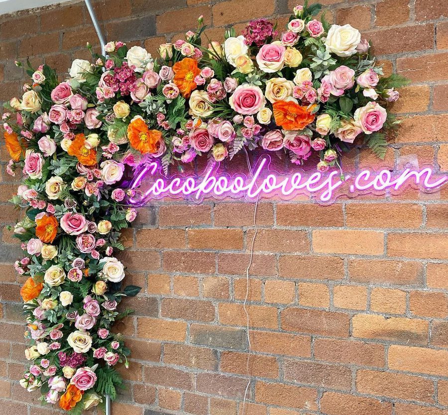 A floral arrangement with pink, white, and orange flowers on a brick wall, featuring a pink neon sign that reads "locopooloves.com".