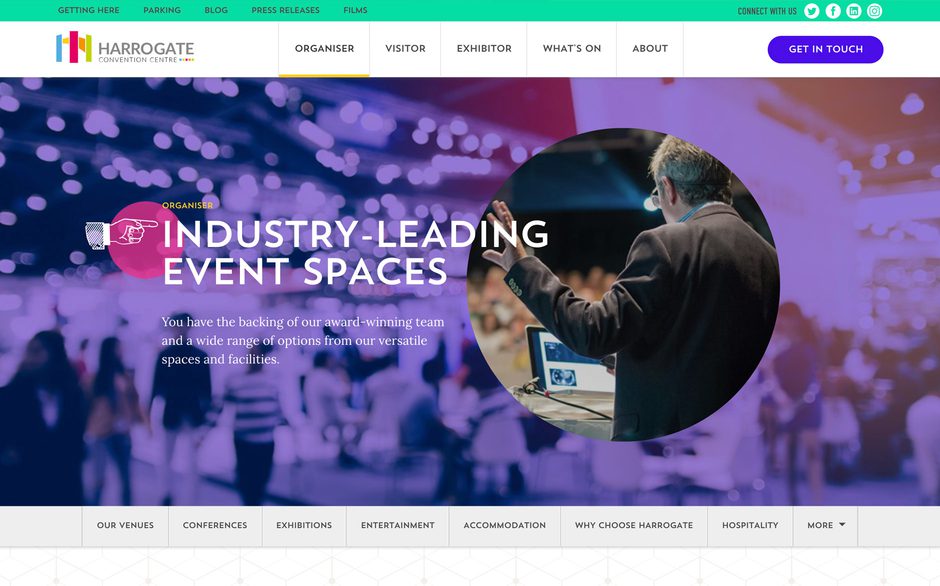 A man in suit gesturing while speaking at a conference, with a blurred audience and colourful bokeh lighting in the background.