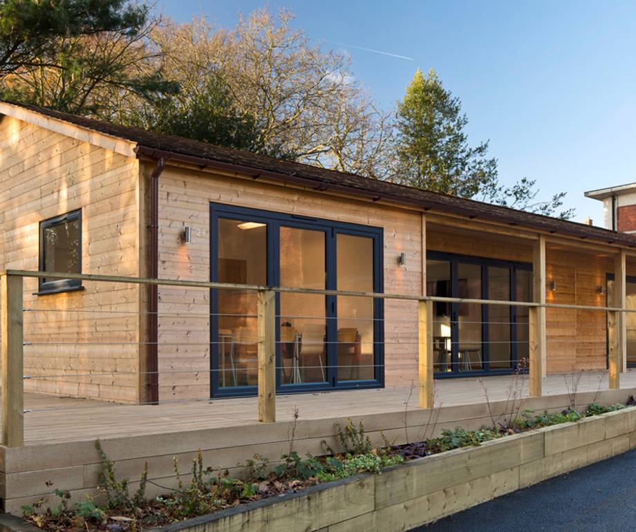 A modern wooden house with large blue-framed glass doors and windows, a wooden balcony with railing, set against a backdrop of trees and a clear blue sky.