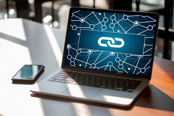 Laptop with a chain link symbol on the screen, surrounded by network graphics, on a wooden table next to a smartphone.