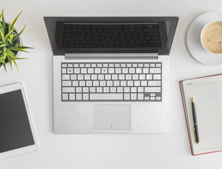 Open laptop, smartphone, cup of coffee, notebook with pen, and a plant arranged on a white desk.