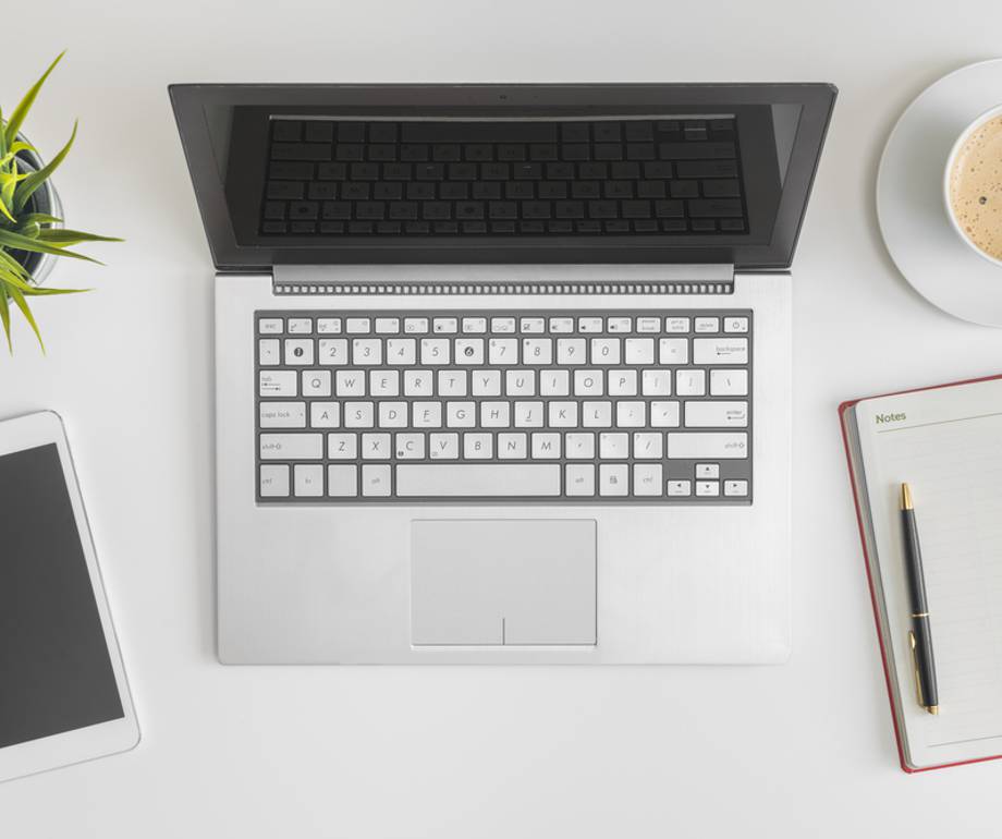 Open laptop, smartphone, cup of coffee, notebook with pen, and a plant arranged on a white desk.