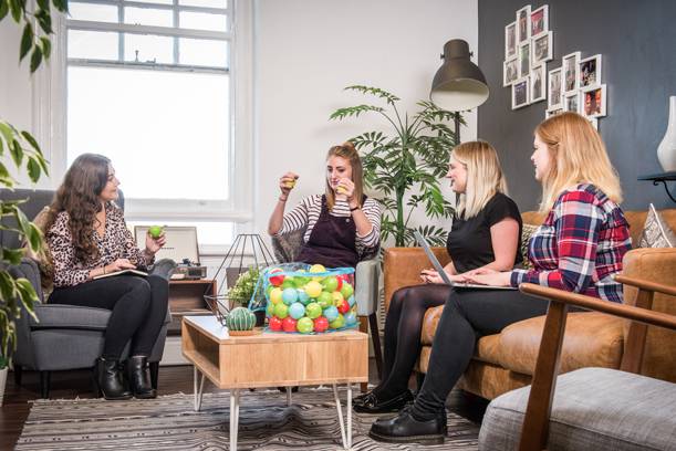 Four women are sitting and chatting in a cosy living room, with a large jar of colourful gumballs on the coffee table.
