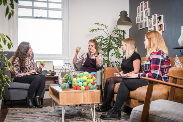 Four women are sitting and chatting in a cosy living room, with a large jar of colourful gumballs on the coffee table.