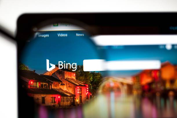 A computer screen displaying the Bing homepage viewed through a magnifying glass, with a traditional building and colourful lights reflected in the glass.