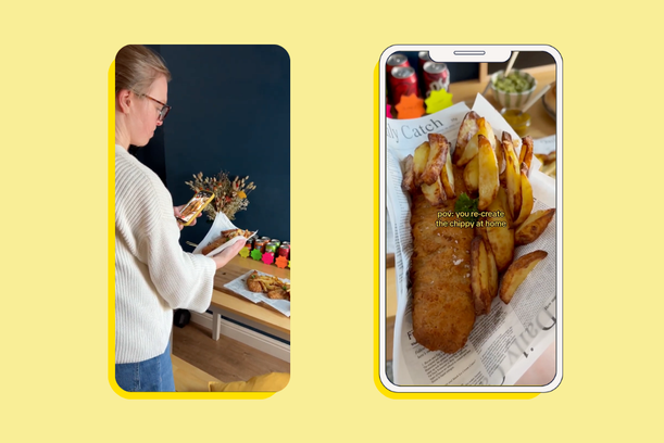 A woman looks at her phone while standing at a table with food, with a phone displaying fried fish and chips in the foreground.