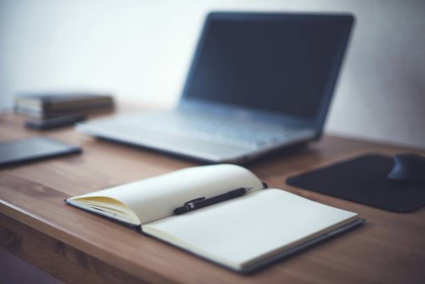 Open notebook with a pen on a wooden desk, with a laptop, mouse and other items blurred in the background.