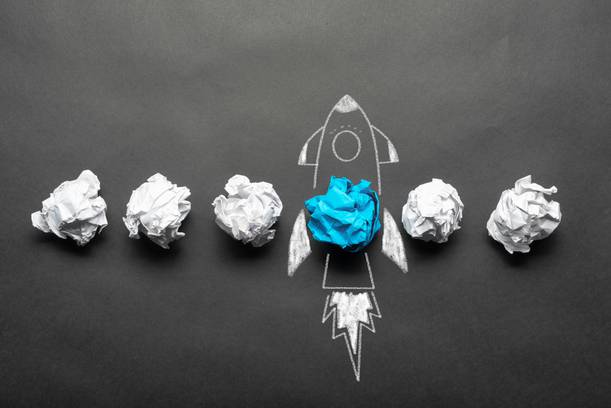 Six crumpled paper balls arranged in a line, with one crumpled blue paper ball in the centre forming part of a chalk-drawn rocket on a dark background.