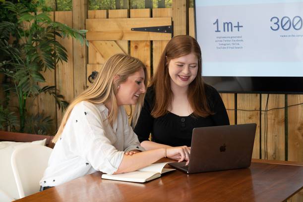 Two women smiling and working together on a laptop at a wooden table in a casual, modern office with a plant and a screen displaying statistics in the background.