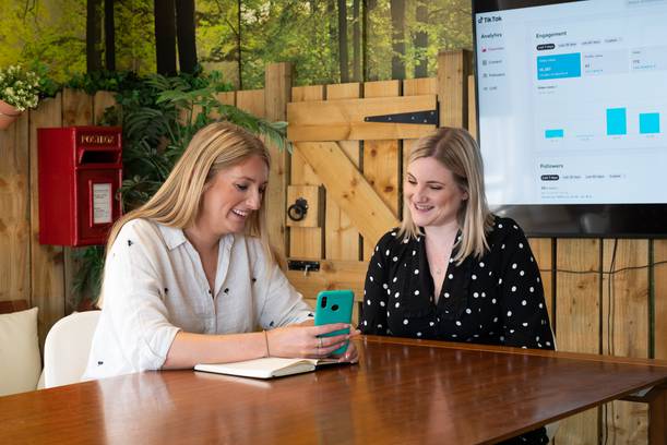 Two women sitting at a wooden table, looking at a phone together, smiling, with a notebook open in front of them, and a large screen displaying TikTok analytics behind them.