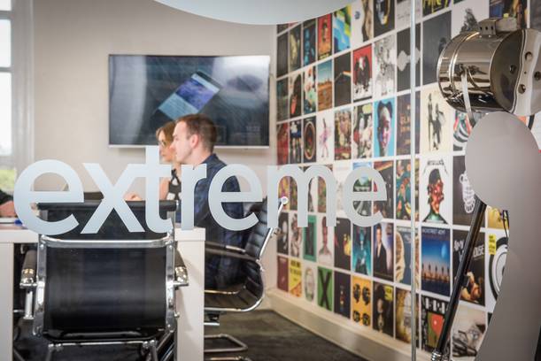 A glass door with the word "extreme" in large letters, showing a meeting room behind with two people seated at a table and a colourful wall of posters.