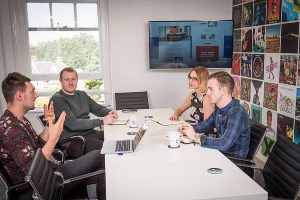 Four people are seated around a white table in a meeting room, engaging in a discussion with notebooks, laptops, and coffee mugs present.