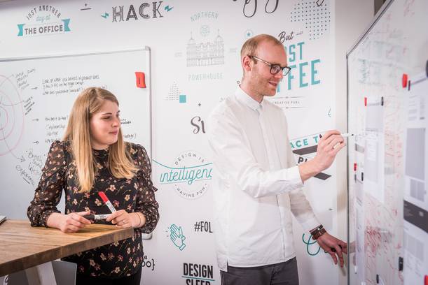 Two people in an office, one woman holding a marker and listening, and a man writing on a whiteboard with notes, in front of a wall with creative designs.