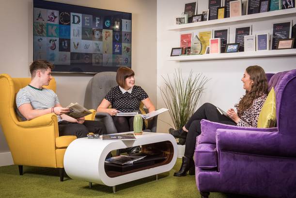 Three women and one man seated on colourful armchairs in a cosy lounge, engaging in a lively discussion with a TV screen displaying alphabet letters in the background.