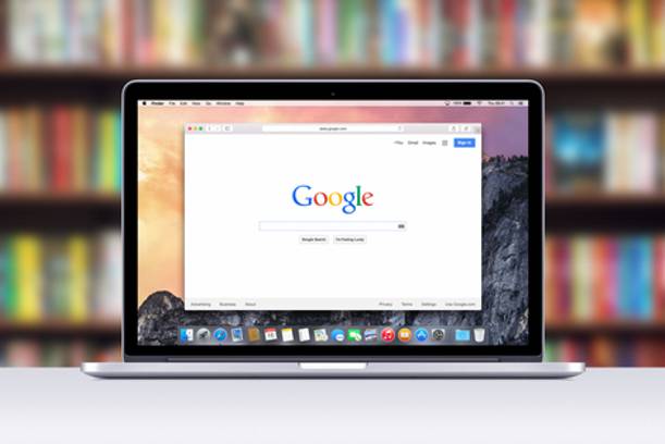 A laptop on a table displays the Google search homepage, with a blurred bookshelf in the background.