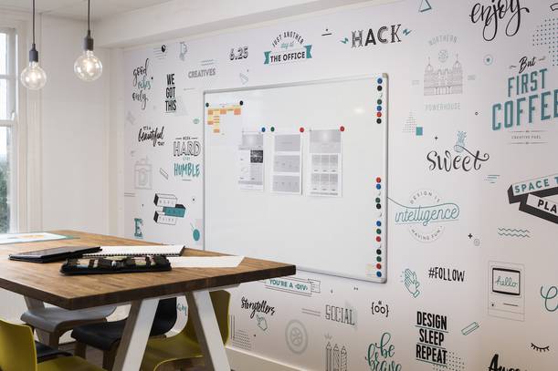 A modern office with a whiteboard on a wall decorated with creative doodles and motivational quotes, alongside a wooden table and chairs.