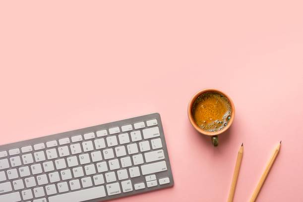 A computer keyboard, two pencils, and a mug of coffee on a pink background.