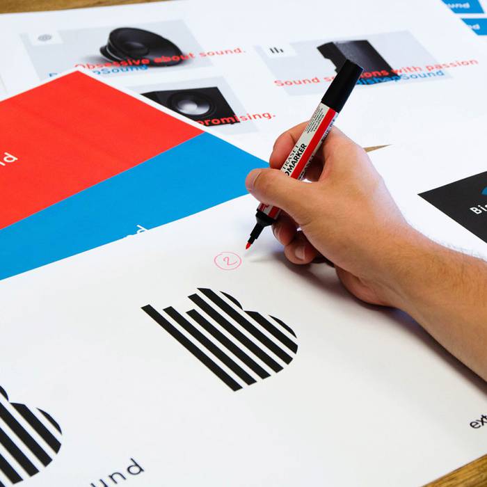 A hand holding a black marker writes on a sheet with black and white striped house illustrations, surrounded by colourful printed pages.