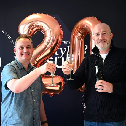 Two men celebrating with glasses of champagne in front of large metallic "20" balloons at a party or event.