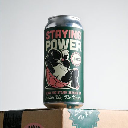 A can of Staying Power IPA with a cartoon turtle illustration, placed on a cardboard box against a plain background.