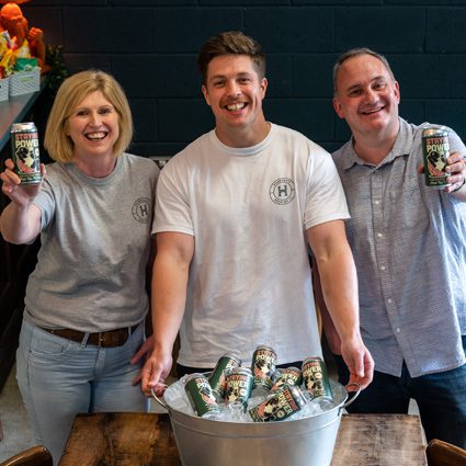 Three smiling people holding cans of drink, standing behind a table and a bucket of cans, in a casual indoor setting with a dark brick wall background.