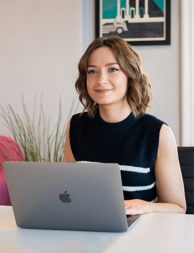 Woman with short wavy brown hair sitting at a desk using a silver MacBook, smiling softly, in a bright, modern room with framed artwork and a plant.