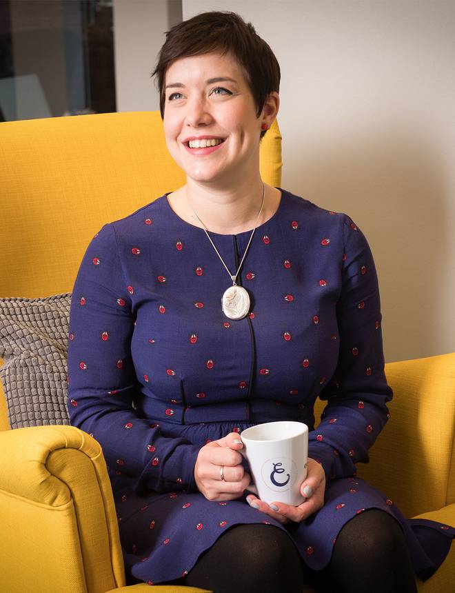 Woman with short dark hair smiling, sitting on a yellow armchair, holding a mug, wearing a navy blue dress with red patterns and a large pendant necklace.