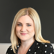 A blonde woman with blue eyes, smiling gently, wearing a black polka dot top, against a blurred dark background.