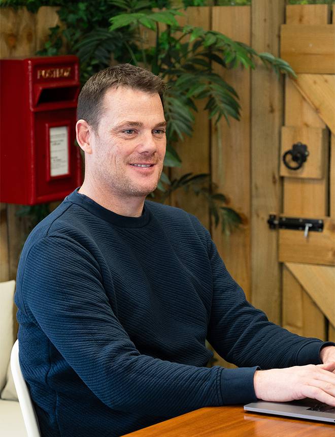 A smiling man in a navy sweater sitting at a table with a laptop, in a wooden outdoor setting with plants and a red fire alarm box in the background.