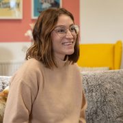 A smiling woman with shoulder-length brown hair, glasses, and a beige turtleneck sweater, sitting on a textured grey sofa in a colourful room.