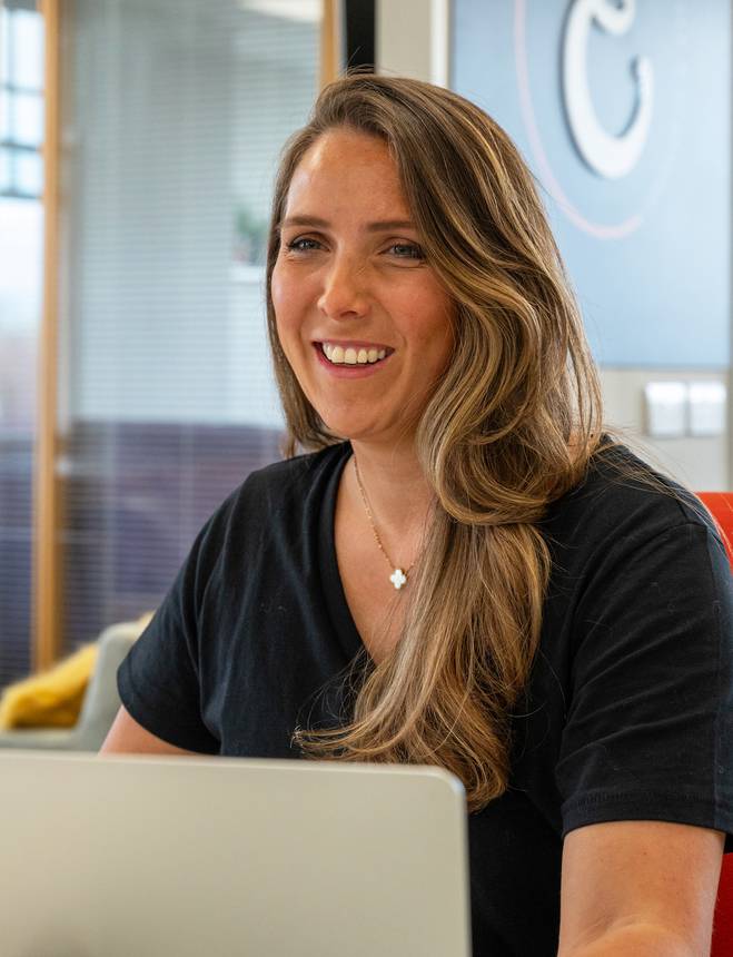 A smiling woman with long wavy hair, wearing a black shirt and a four-leaf clover necklace, sitting in front of a laptop in an office setting.