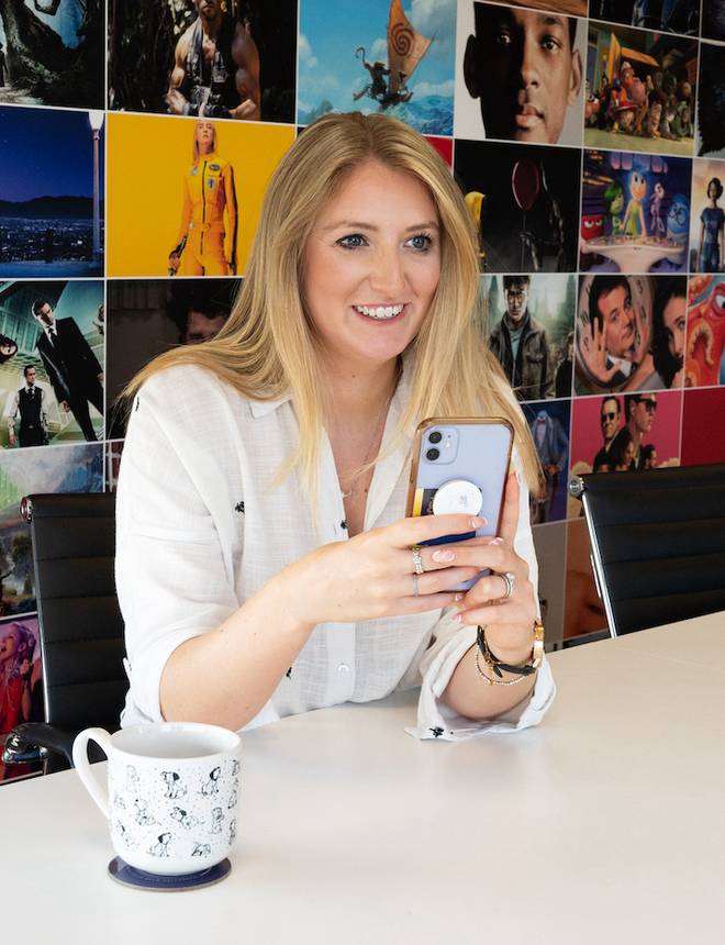 Woman with long blonde hair, smiling and looking at her phone, seated at a table with a mug, with a colourful collage of animated and live-action images behind her.