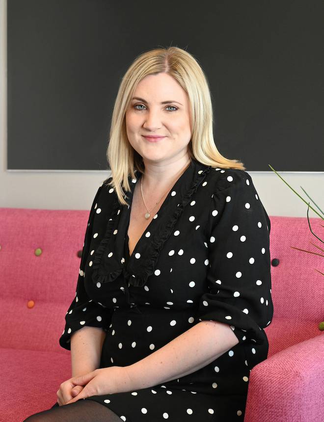 Woman with blonde hair sitting on a pink sofa, wearing a black polka dot dress and smiling, in front of a dark grey wall.