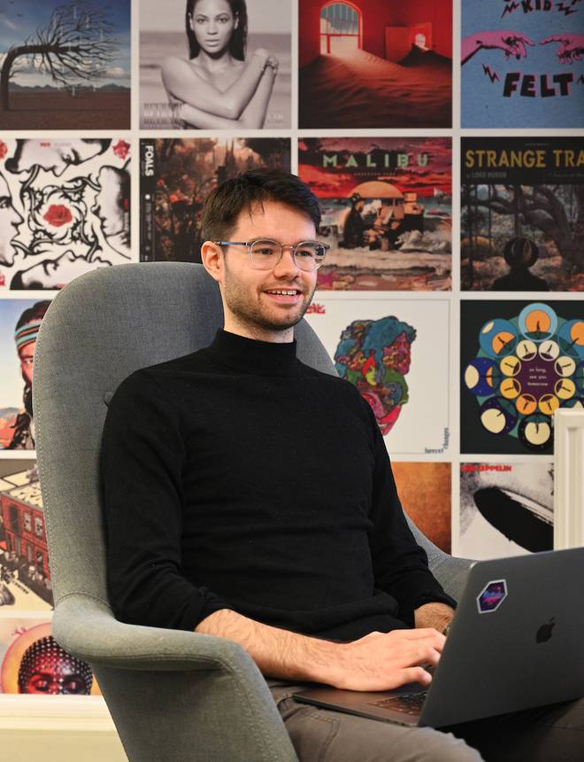 A young man with glasses, wearing a black turtleneck, sitting in a grey armchair with a colourful album covers wall in the background, working on a laptop.