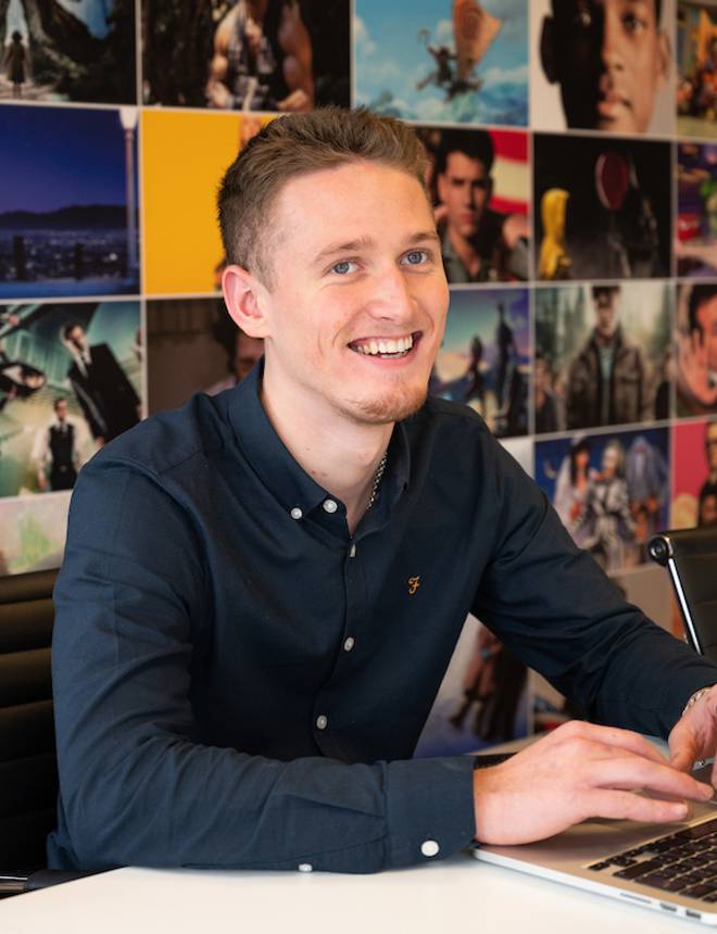 Young man with short hair in a navy shirt, smiling while working on a laptop in a vibrant office with a colourful photo collage background.