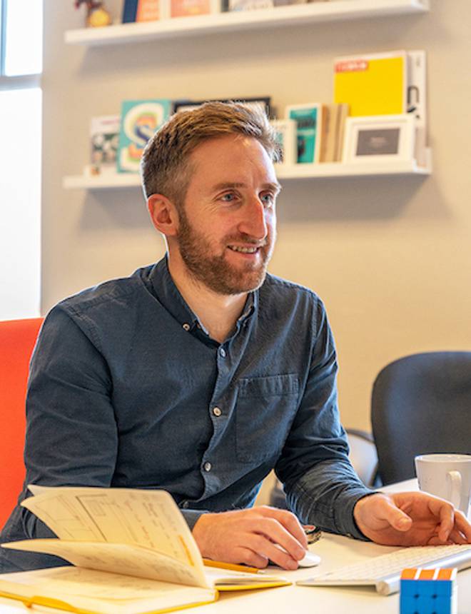 A man with short brown hair and a beard, wearing a dark blue shirt, sitting at a table with open books, in a room with bookshelves behind.