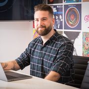 A young man with a beard and checked shirt sitting at a desk with a laptop, smiling against a colourful poster-filled wall.