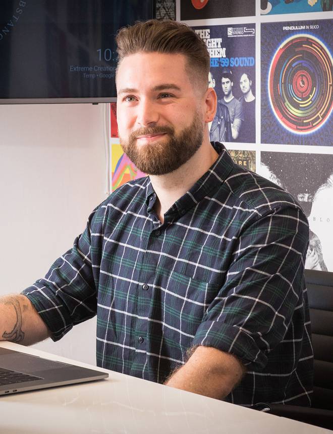 A young man with a beard and checked shirt sitting at a desk with a laptop, smiling against a colourful poster-filled wall.
