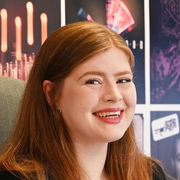 A young woman with long red hair smiling in front of a colourful background with abstract images and patterns.