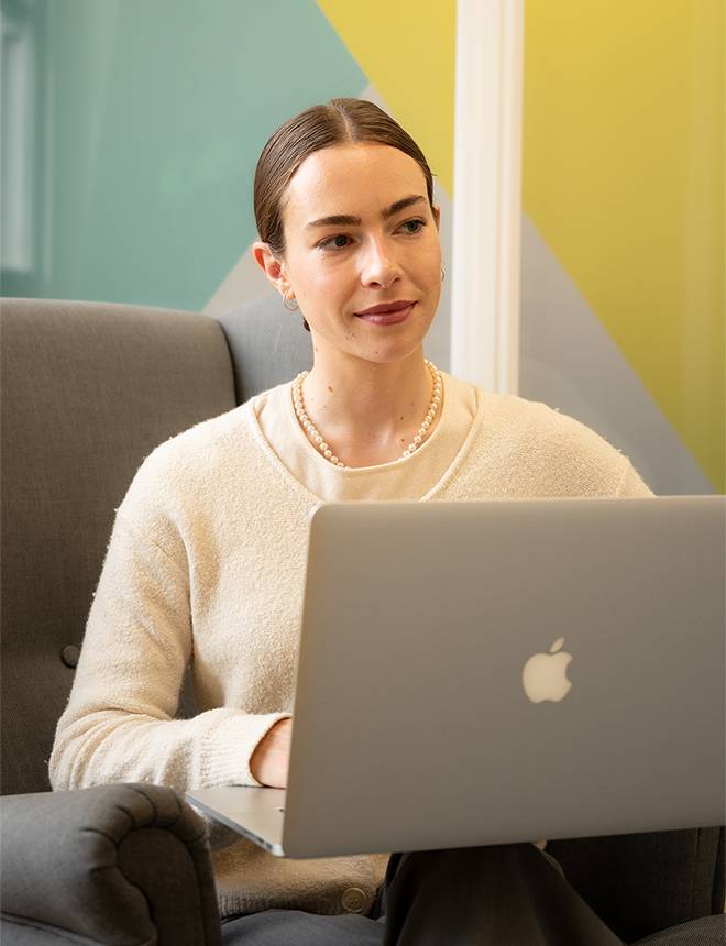 A woman with brown hair and pearl necklace working on a silver Apple MacBook in a colourful modern interior.