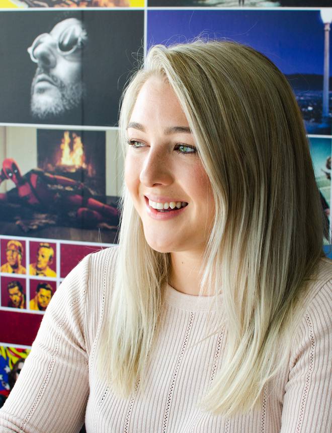A smiling blonde woman sitting in front of a collage of colourful images on the wall, with blue chairs partly visible in the foreground.