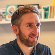 A smiling man with short hair and a beard, sitting indoors with shelves of books and artwork in the background.