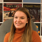Young woman with long hair smiling, sitting in front of a wall decorated with travel posters.