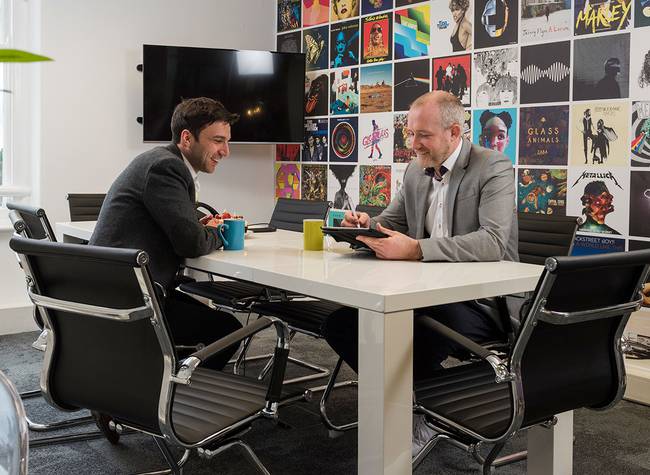 Two men in business attire sit across from each other at a white conference table, smiling and engaging in conversation in a modern office.