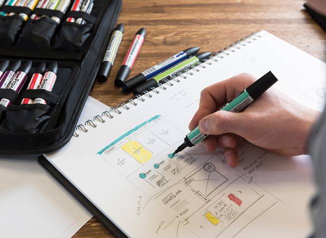 Person sketching a website layout in a notebook, surrounded by colourful markers and a case on a wooden table.
