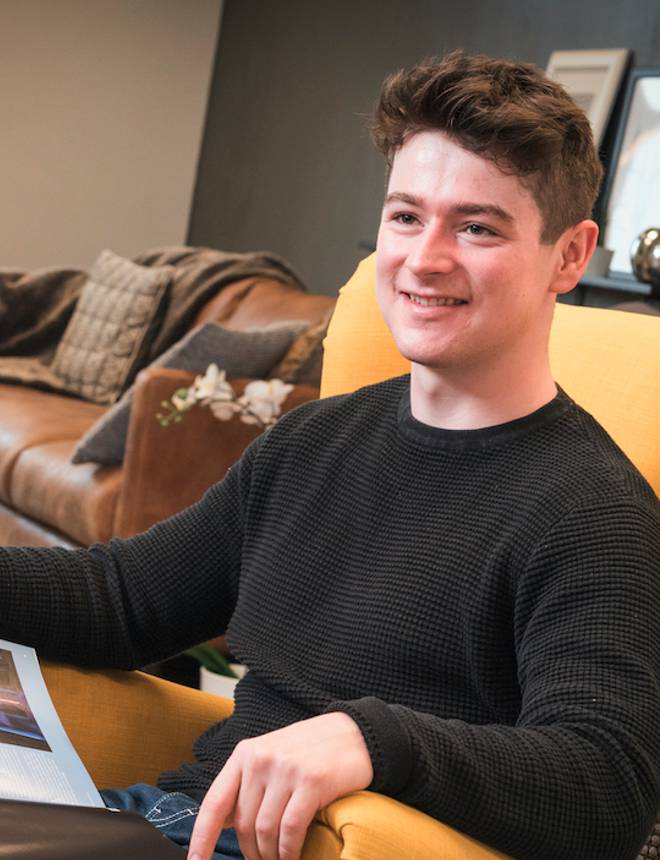 Young man sitting on a yellow armchair, smiling, with a magazine on his lap in a cozy living room setting.