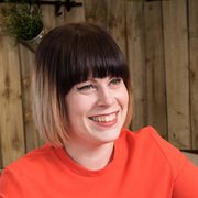 Woman with short dark brown hair and blunt bangs, smiling and wearing an orange top, set against a wooden background.