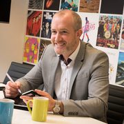 A smiling man in a grey suit sitting at a table with a phone in hand, colourful posters on the wall behind him in an office setting.