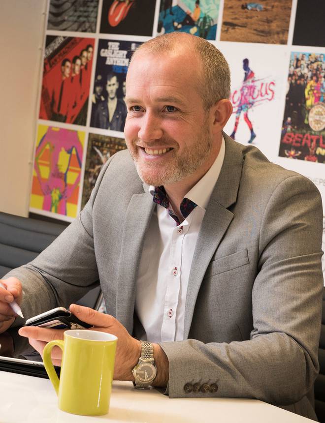 A smiling man in a grey suit sitting at a table with a phone in hand, colourful posters on the wall behind him in an office setting.